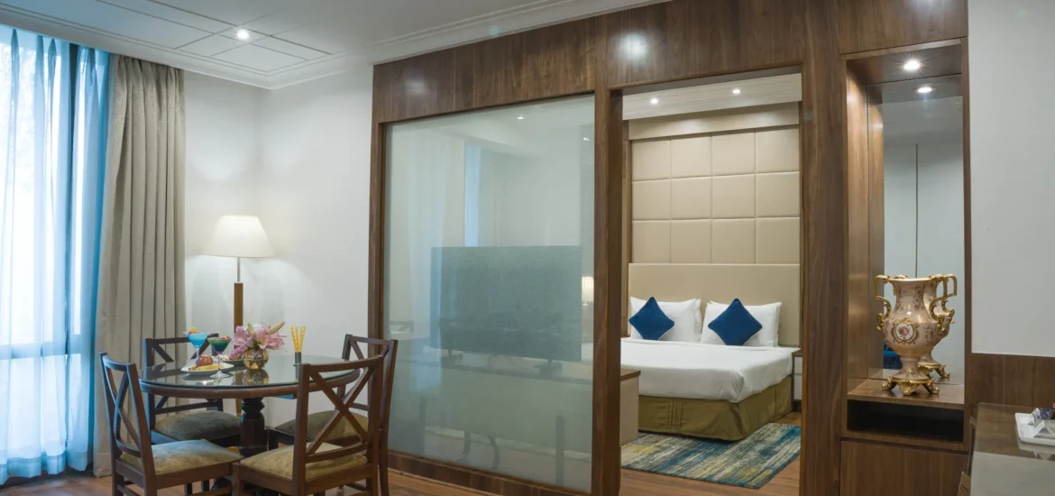 Deluxe Suite Hotel room interior with a dining area, bed with blue cushions, and wooden partition.